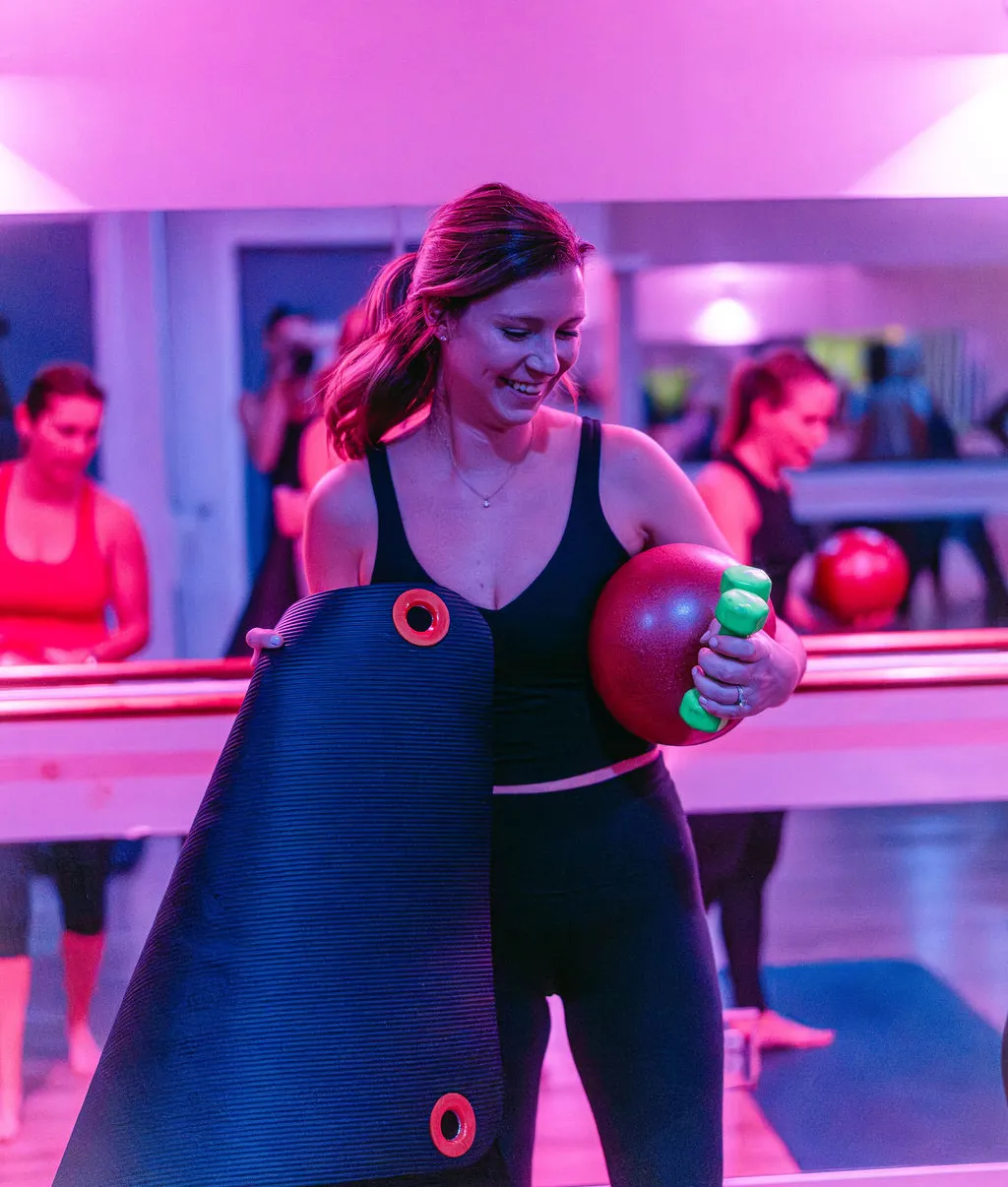 Woman in workout clothes holding a yoga mat, exercise ball, and weights in a pink-lit fitness studio.
