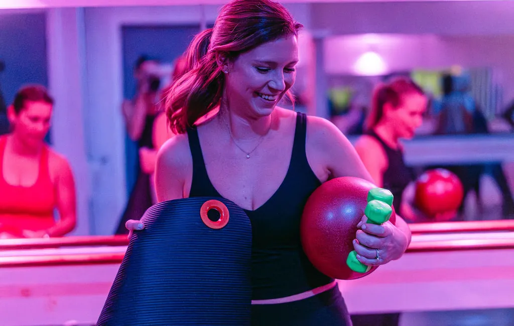 Woman in workout gear holding a yoga mat, red ball, and green dumbbells in a fitness studio with pink lighting.