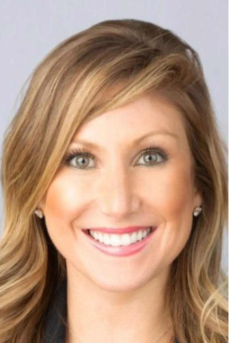 Smiling woman with wavy blonde hair, light eyes, and wearing earrings, pictured against a plain background.