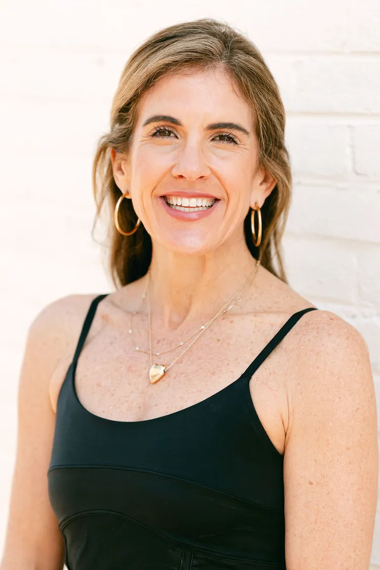 Smiling woman with long hair, hoop earrings, and layered necklaces, wearing a black top, standing by a white wall.