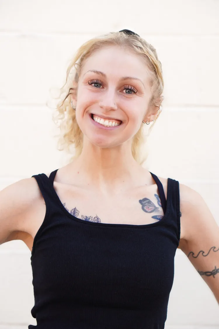 Smiling woman with blonde curly hair in a black tank top, showing tattoos on her arms and chest, against a light wall.