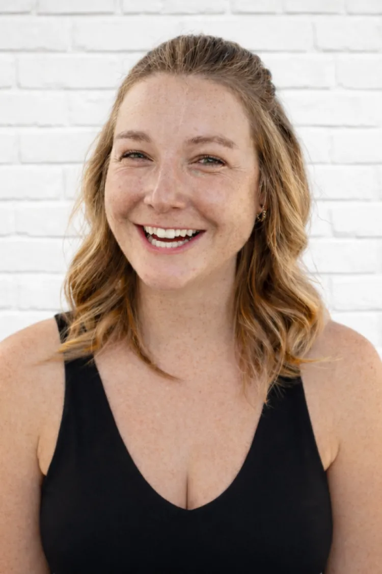Smiling woman with wavy hair wearing a black top, standing in front of a white brick wall.