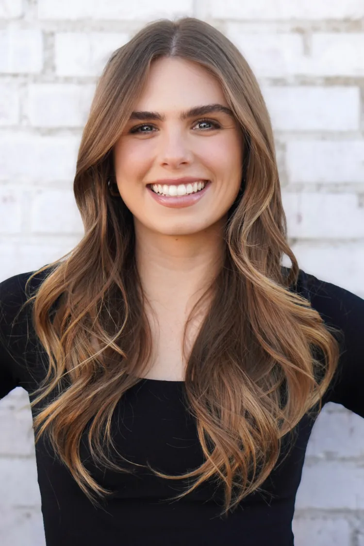 A woman with long wavy brown hair smiles in front of a white brick wall, wearing a black top.