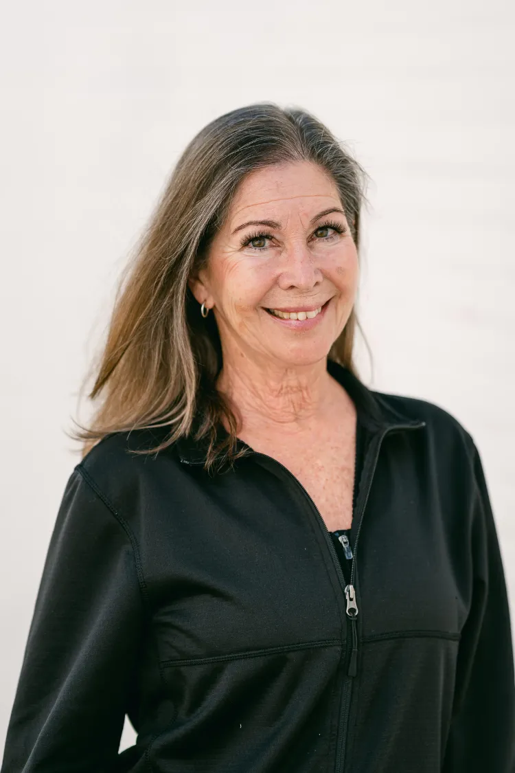 Smiling woman with long brown hair wearing a black zip-up jacket, standing against a light background.