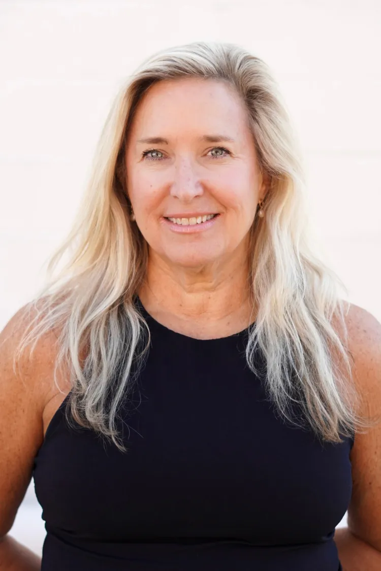 A smiling woman with long blonde hair wearing a sleeveless black top against a light background.