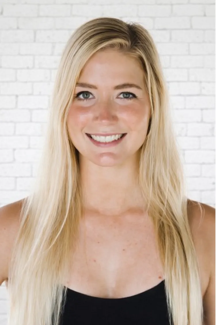 Smiling woman with long blonde hair in a black top standing in front of a white brick wall.
