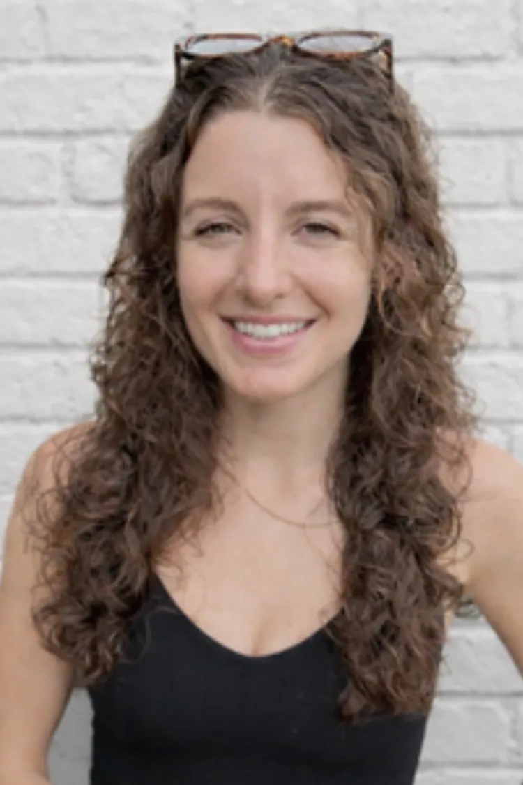 Smiling woman with curly brown hair and glasses on head stands in front of a white brick wall.