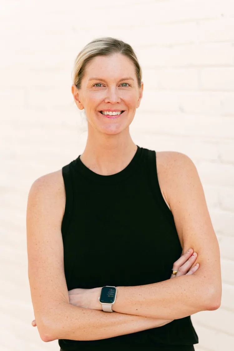 Smiling woman in a black sleeveless top with arms crossed, wearing a smartwatch, standing against a white wall.