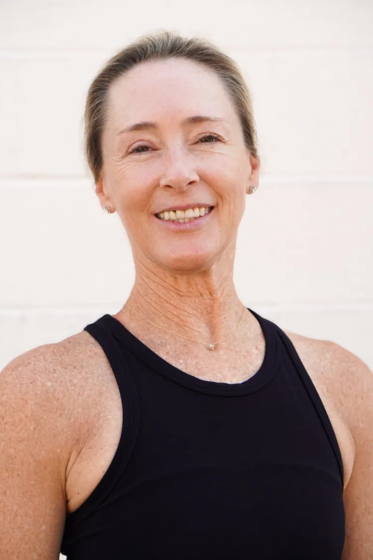 Smiling woman with light hair in a black tank top standing in front of a light-colored wall.