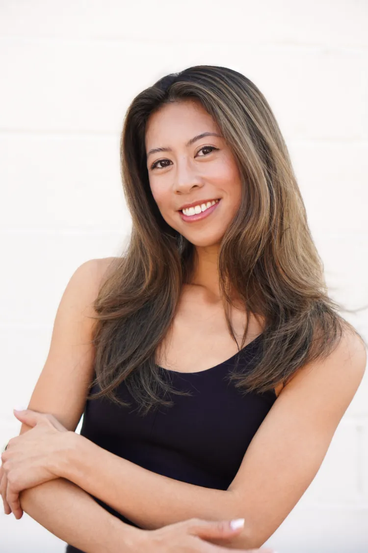 Smiling woman with long brown hair stands against a white background, arms crossed, wearing a black top.