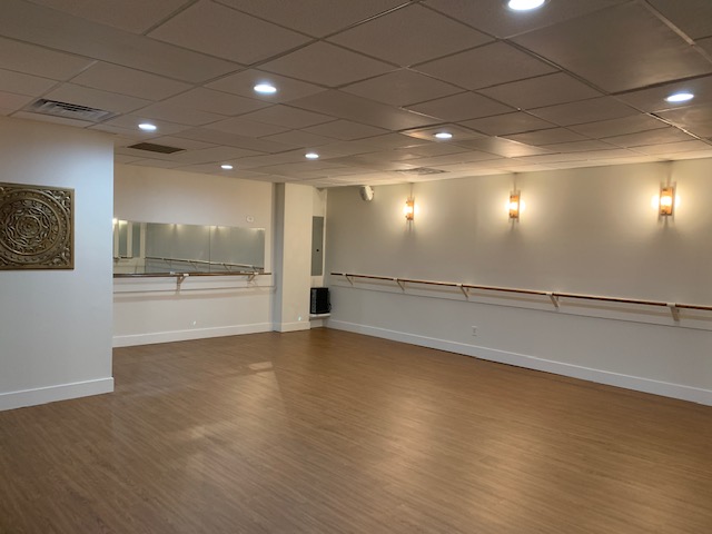IMG_4750 Spacious dance studio with wooden floor, wall mirrors, ballet barres, and soft lighting on white walls.