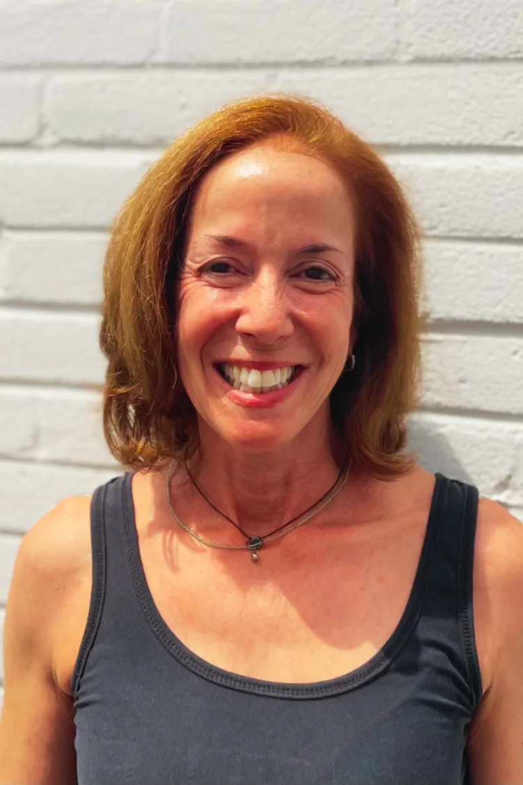 Smiling woman with shoulder-length brown hair wearing a dark tank top, standing in front of a white brick wall.