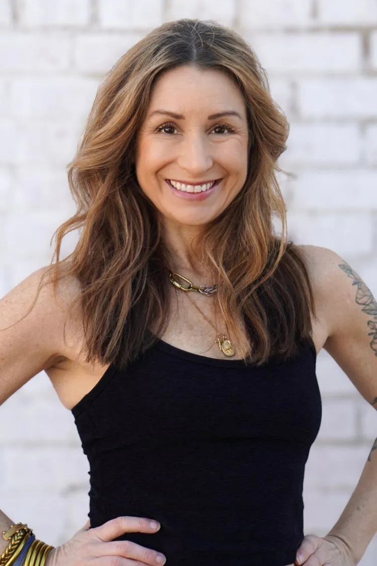 Smiling woman with long brown hair in a black tank top, standing in front of a white brick wall.