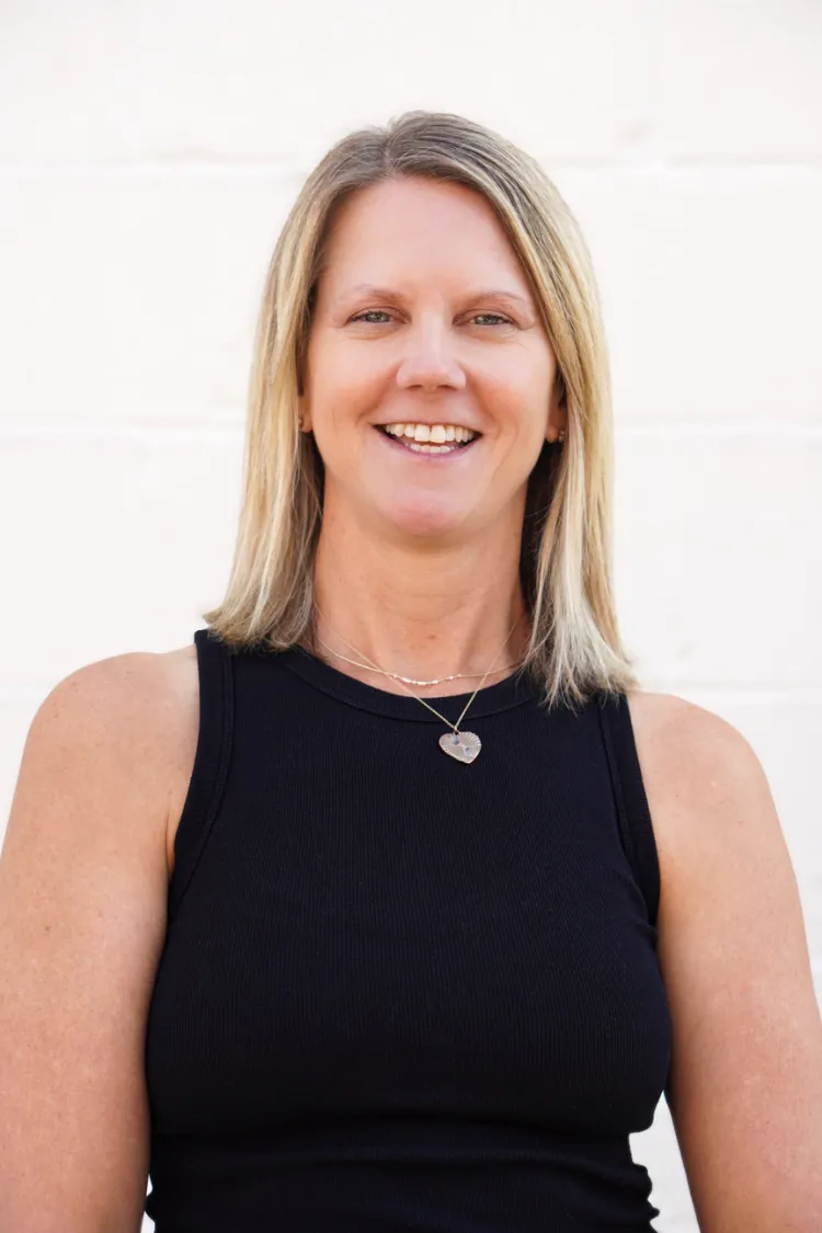 Smiling woman with straight blonde hair wearing a sleeveless black top and a heart-shaped necklace.