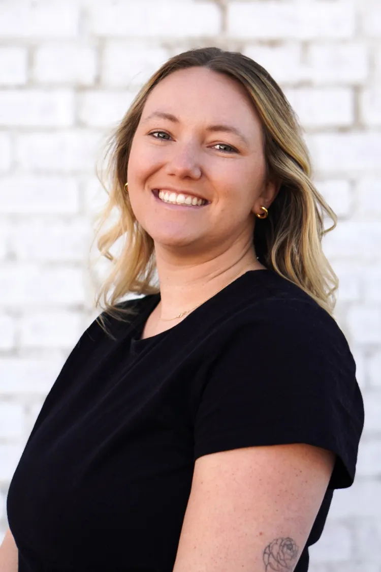 Woman with blonde hair in a black shirt smiling in front of a white brick wall.