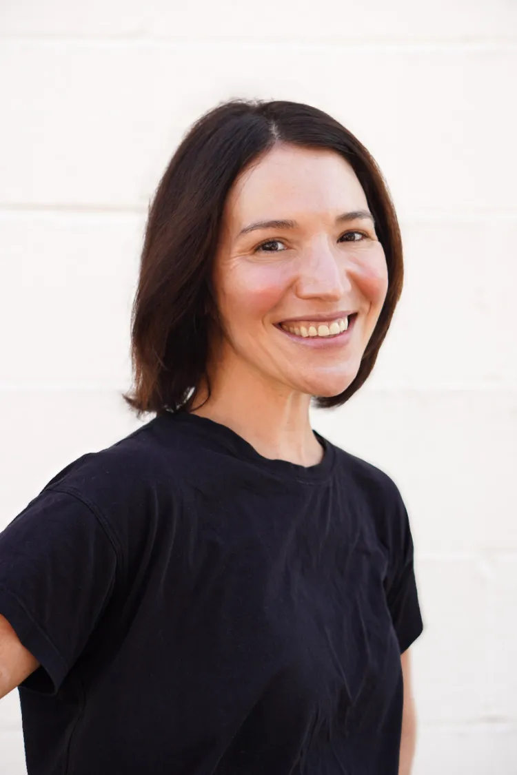 Woman with shoulder-length brown hair smiling, wearing a black t-shirt, standing against a white background.