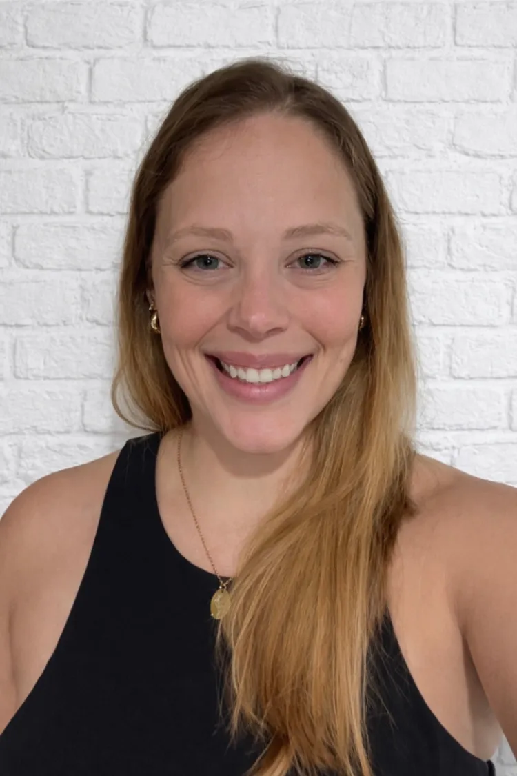 Woman with long blonde hair in a black top smiling in front of a white brick wall.