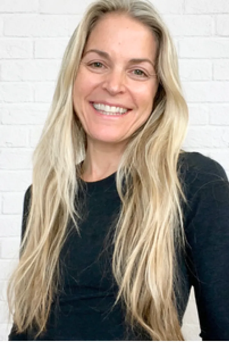 Smiling woman with long blonde hair wearing a black top, standing in front of a white brick wall.