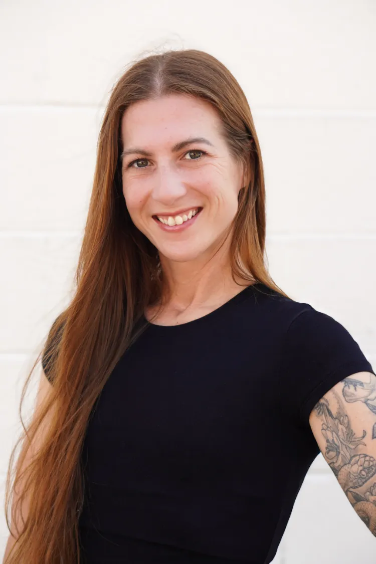 Smiling woman with long brown hair, wearing a black shirt, shows a tattoo on her arm, standing against a light wall.