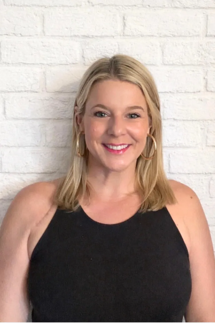 Woman with blonde hair in a black sleeveless top, smiling in front of a white brick wall.