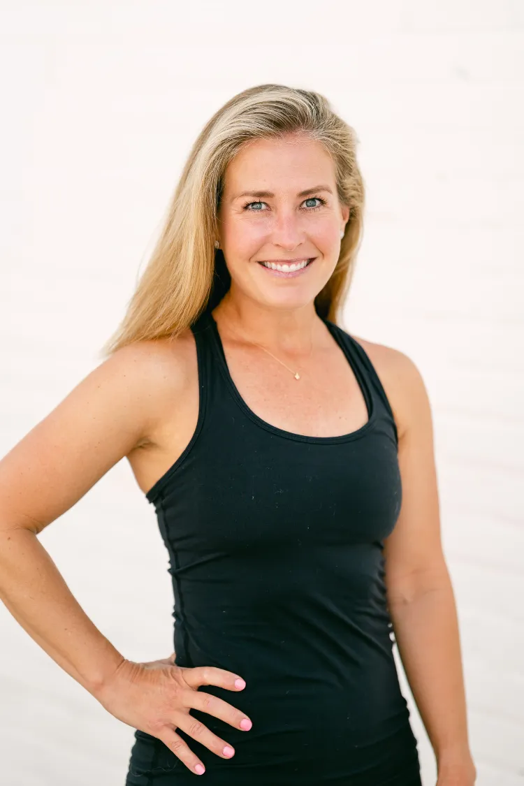 Woman with long blonde hair wearing a black tank top, smiling with one hand on her hip against a light background.