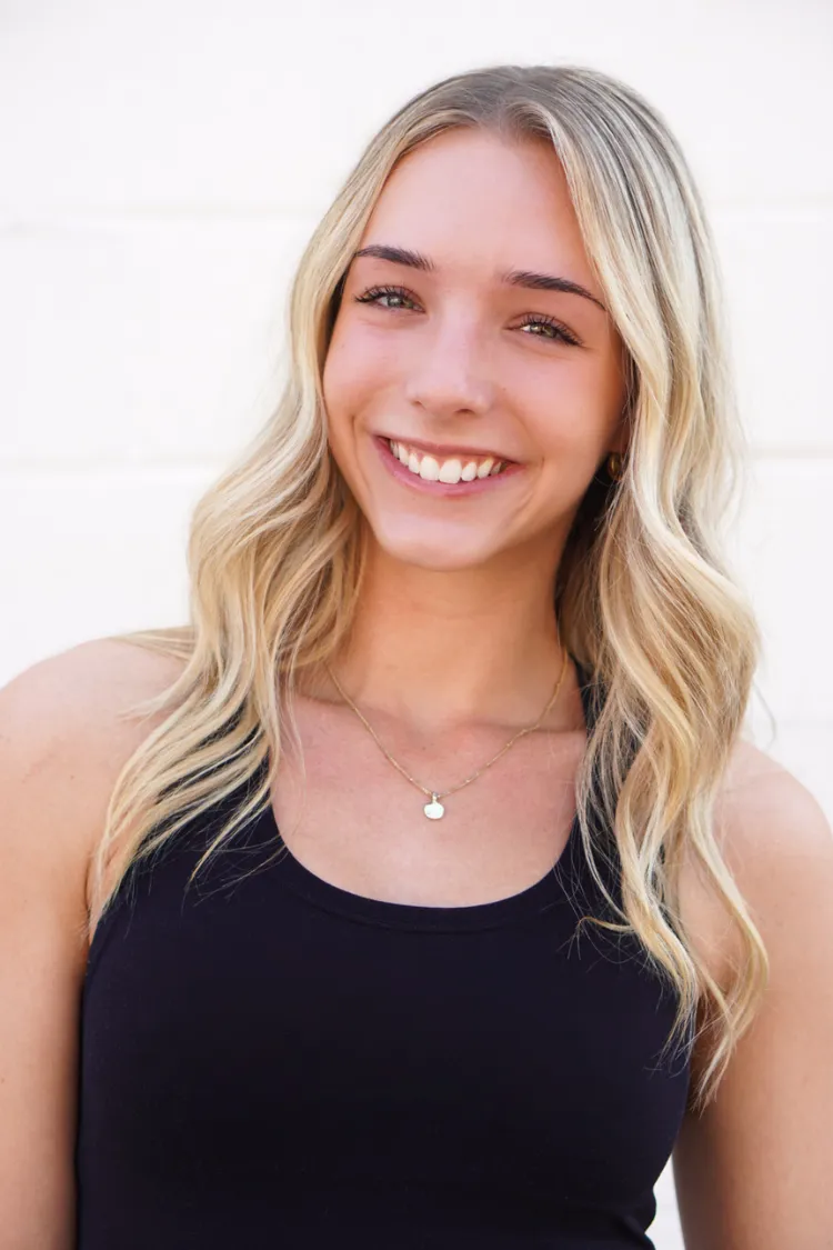 Young woman with long blonde hair in a black tank top, smiling in front of a white background.