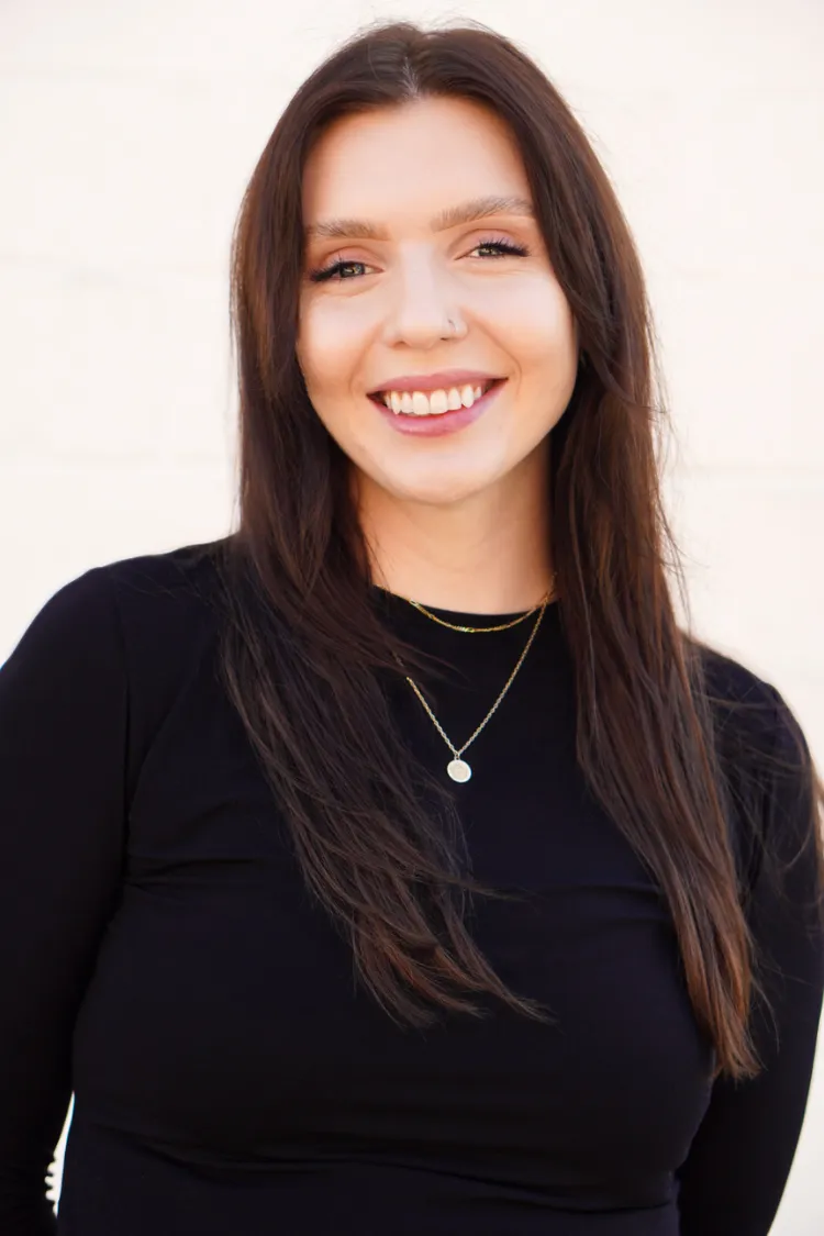 Woman with long brown hair wearing a black top, smiling in front of a light-colored background.