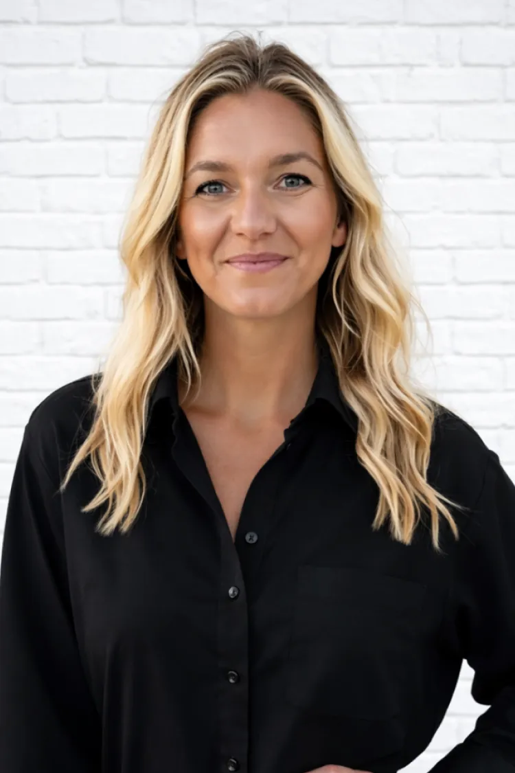 Smiling woman with long blonde hair in a black shirt stands in front of a white brick wall.