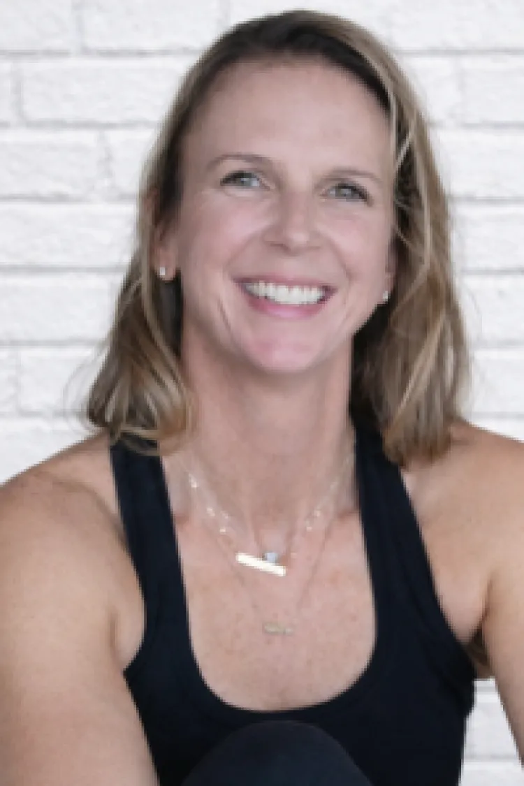 Smiling woman with shoulder-length hair wearing a black tank top, sitting in front of a white brick wall.