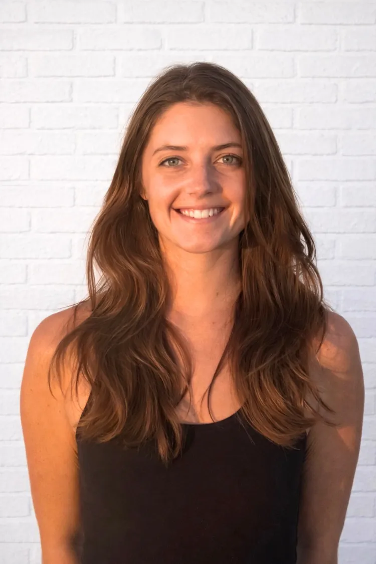 A woman with long brown hair and a black tank top smiles in front of a white brick wall.