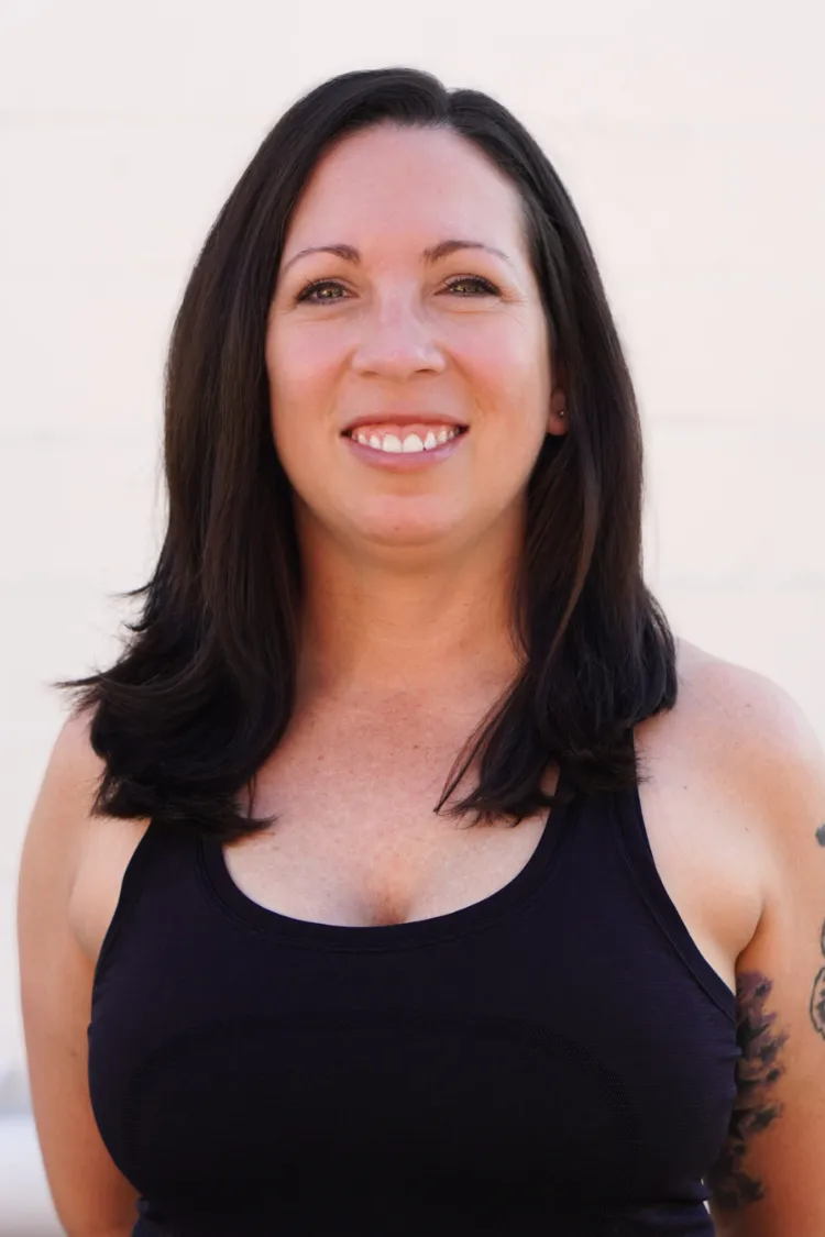Woman with long dark hair smiling, wearing a black tank top, standing against a plain light background.