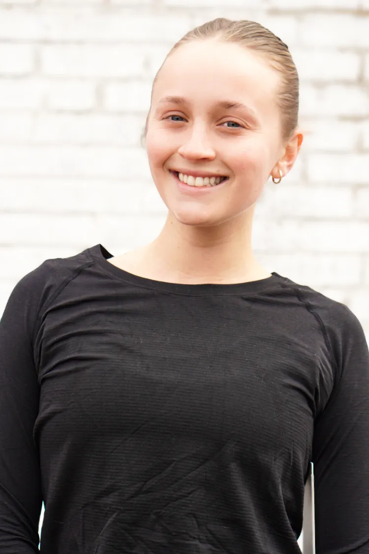 Young woman with light hair in a bun, wearing a black top, smiling in front of a white brick wall.