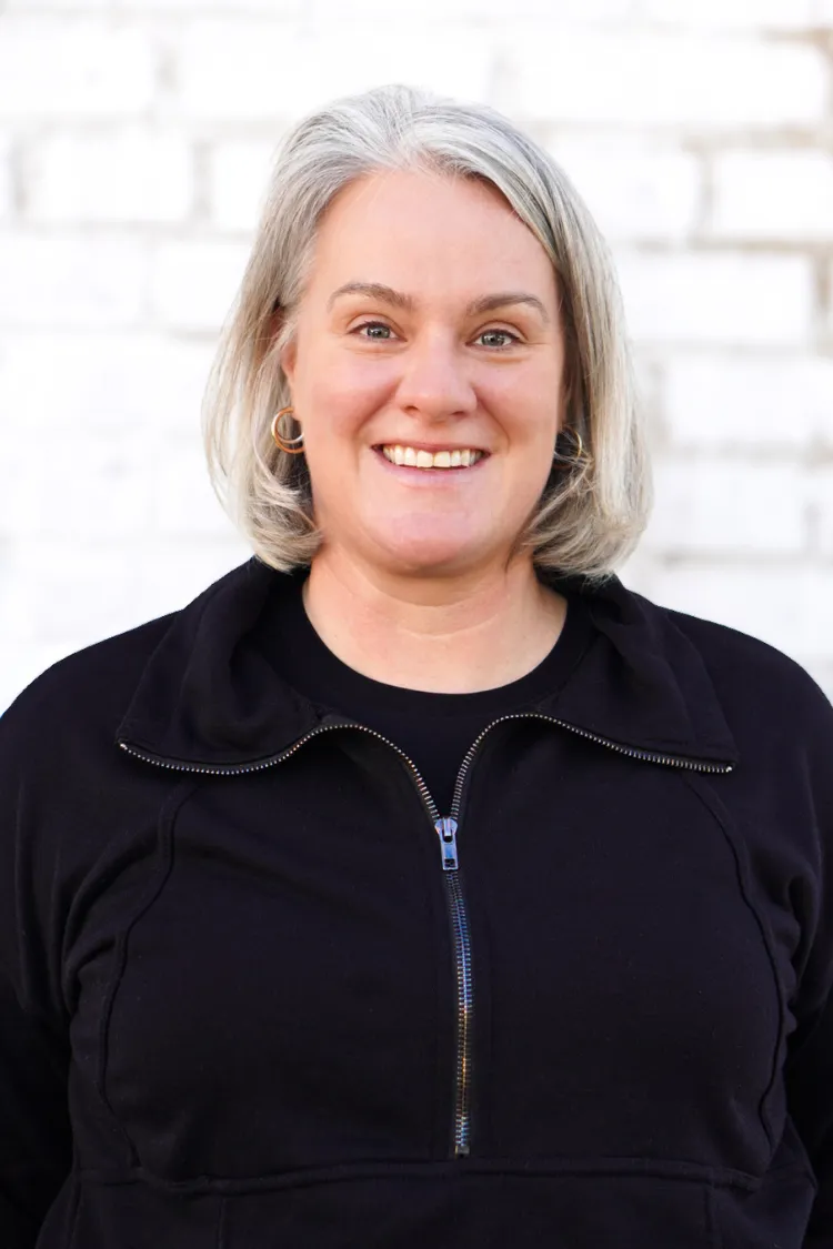 Smiling woman with gray hair wearing a black zip-up jacket stands in front of a white brick wall.