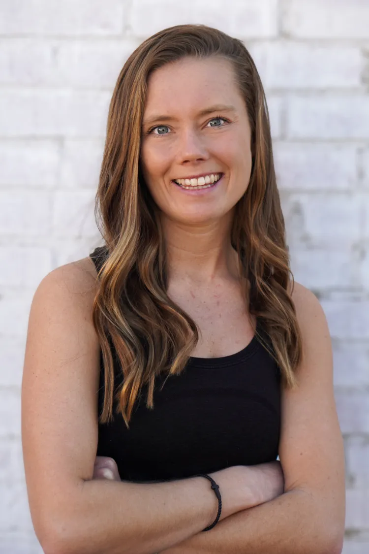 Woman with long brown hair in a black tank top, smiling with arms crossed, standing in front of a white brick wall.