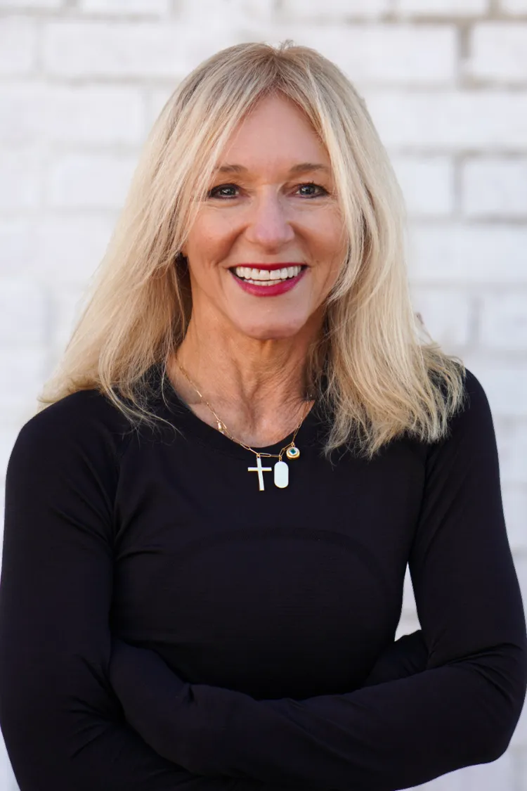 Smiling woman with long blonde hair in a black top, wearing a cross necklace, standing against a white brick wall.