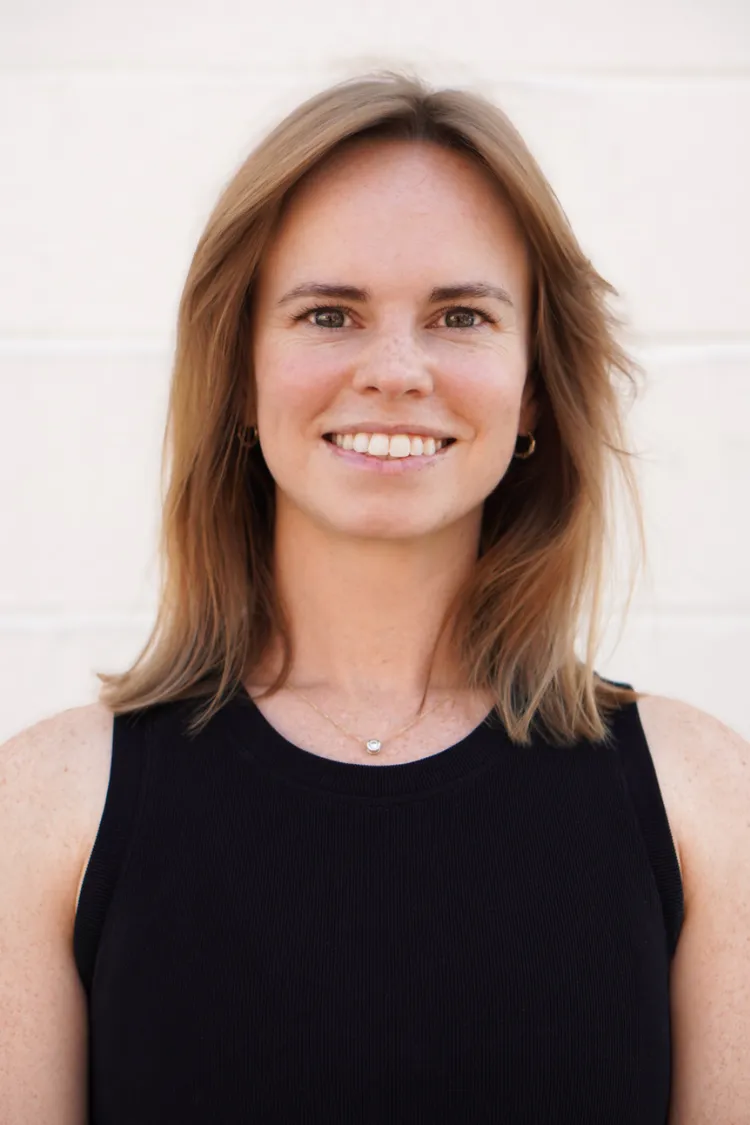 Woman with shoulder-length light brown hair smiling, wearing a black sleeveless top and a necklace, against a light wall.
