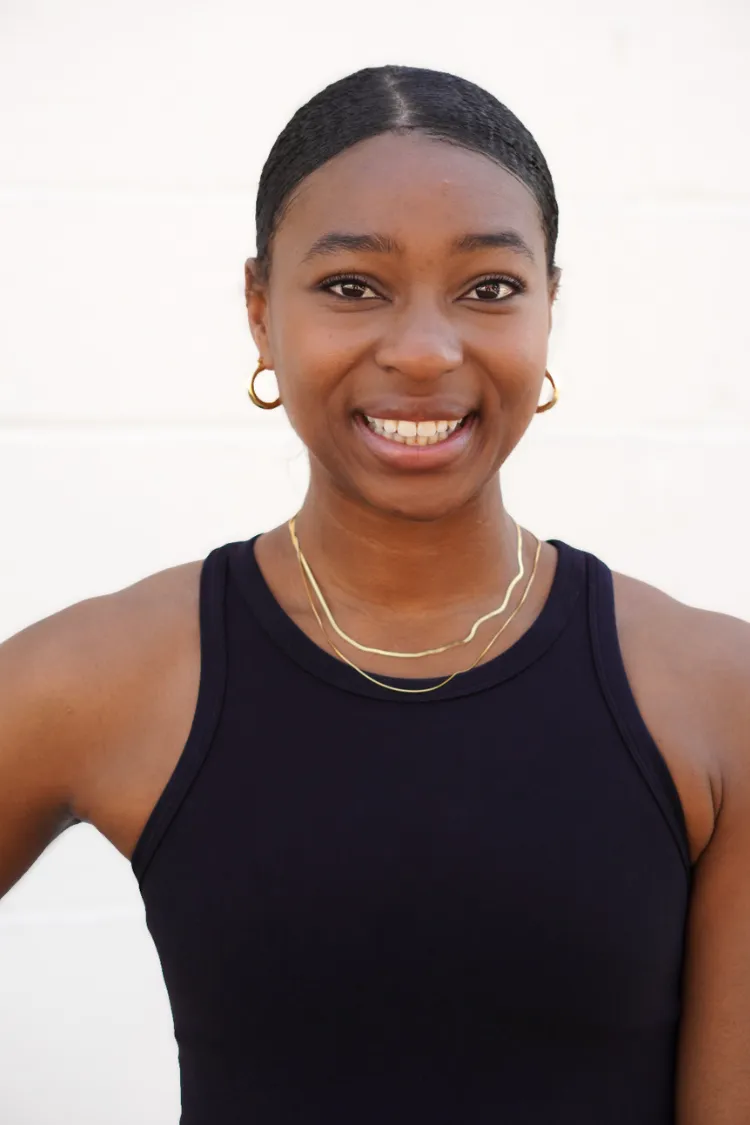 Victoria Norris Smiling woman with gold hoop earrings and necklace, wearing a sleeveless black top, standing against a white background.