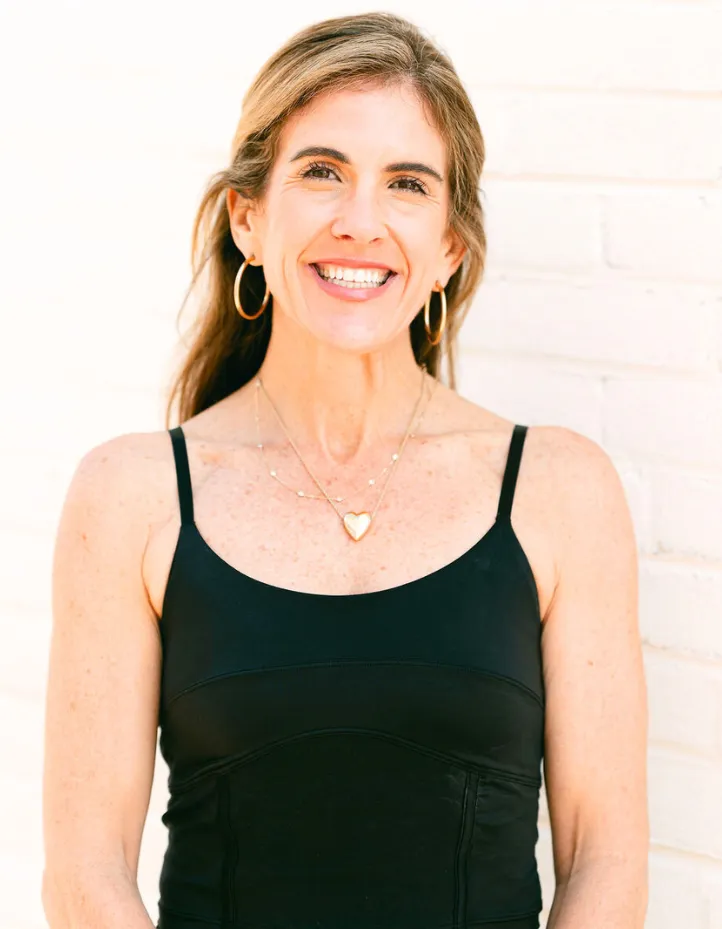 annasegrave-1 Smiling woman with long hair in a black tank top, gold hoop earrings, and layered necklaces, standing by a white wall.