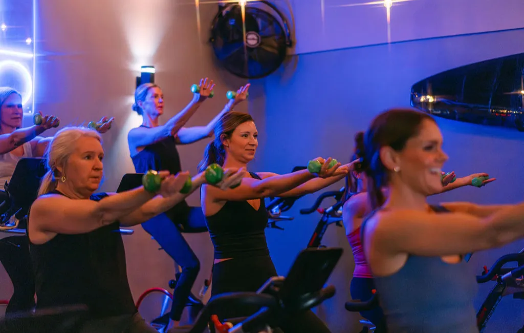 A group of women in a fitness class holds small weights while riding stationary bikes under blue lighting.