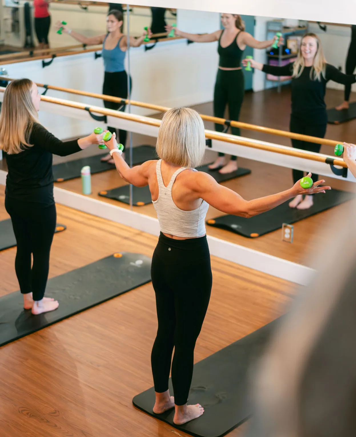 Women in workout clothes hold small weights, exercising in front of mirrors in a fitness studio.