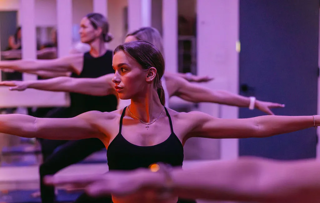 Women in athletic wear practicing yoga in a studio, holding the Warrior II pose with arms extended.