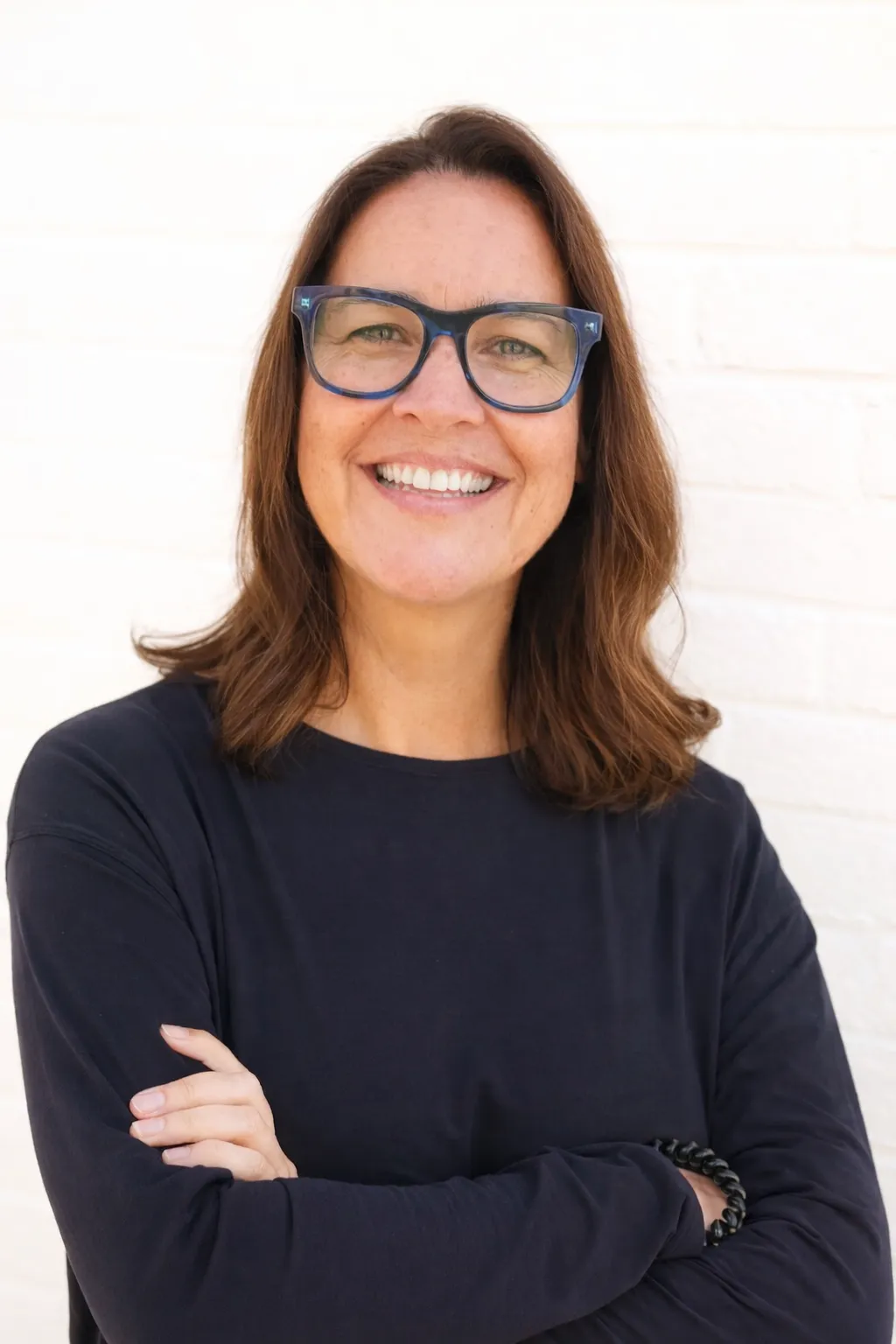sam-light Woman with shoulder-length brown hair, glasses, and a black top smiling with arms crossed against a white background.
