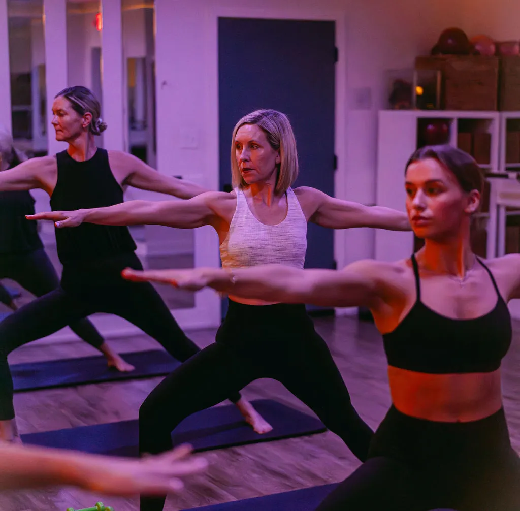 studio-1 Three women in athletic wear practice yoga in a studio, holding Warrior II pose on yoga mats.