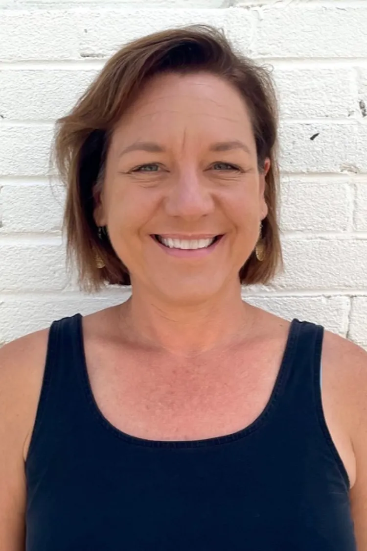Woman with short brown hair, wearing a black tank top, smiling in front of a white brick wall.