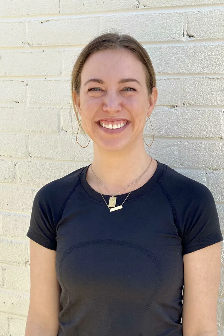 Smiling woman with brown hair in a ponytail, wearing a black shirt, stands against a white brick wall.