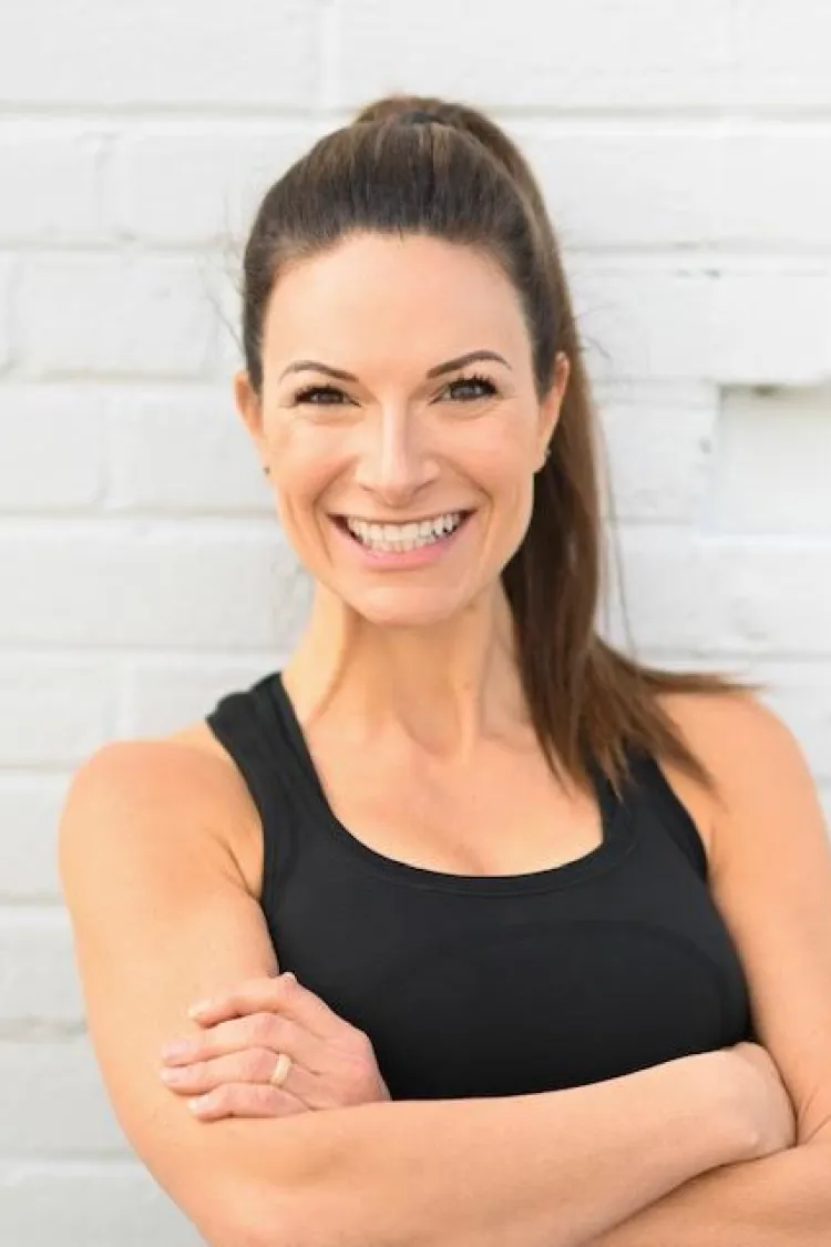 Woman with brown hair in a ponytail, wearing a black tank top, smiles with arms crossed against a white brick wall.