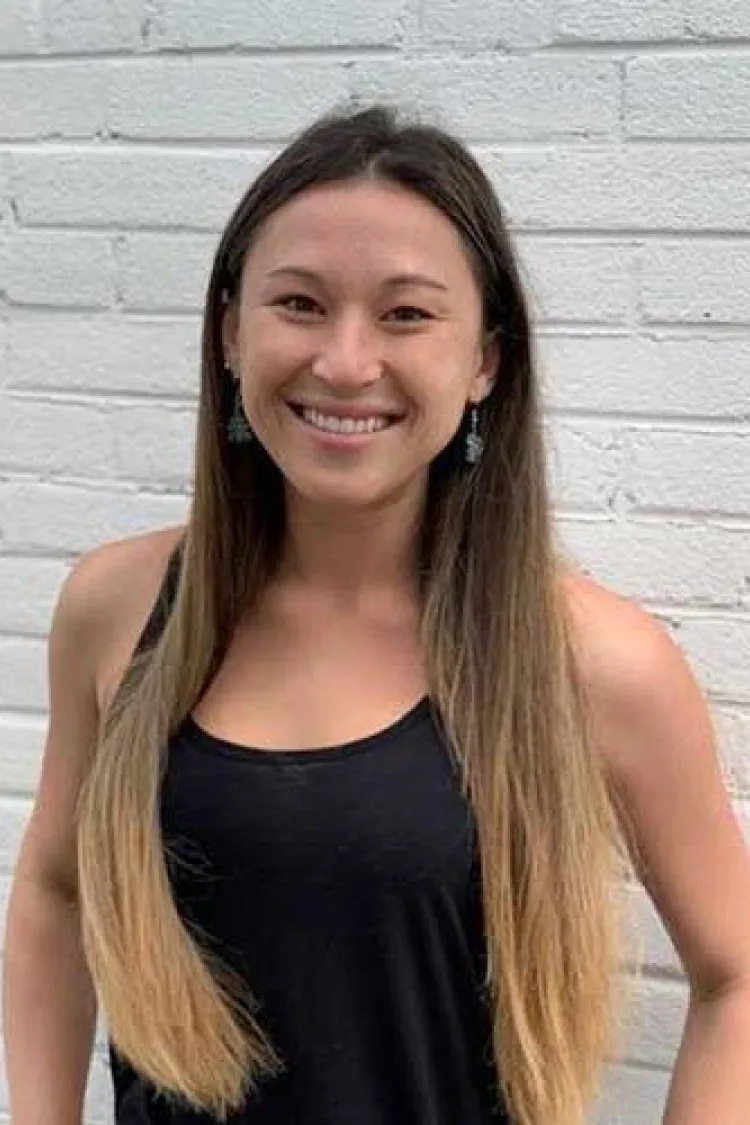 Woman with long, straight brown hair wearing a black tank top stands smiling in front of a white brick wall.