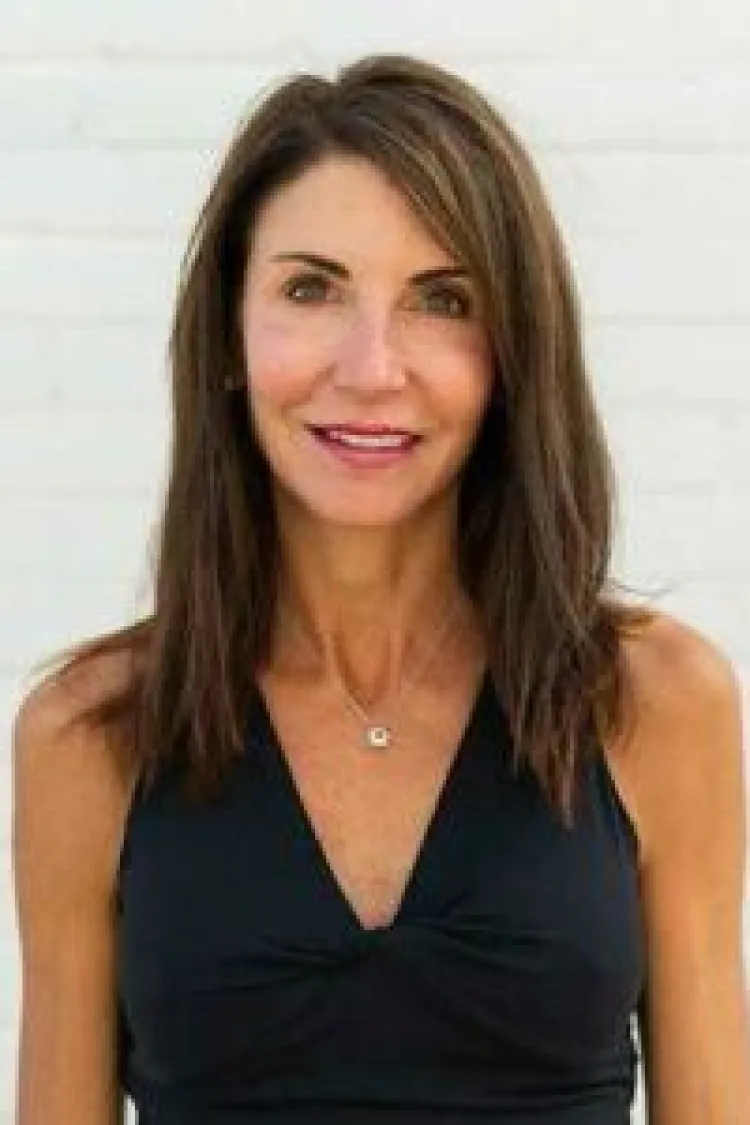 Woman with straight brown hair wearing a sleeveless black top, standing in front of a white background, smiling.