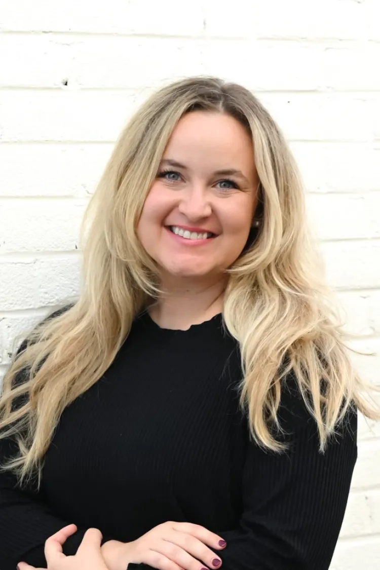 Smiling woman with long blonde hair in a black top standing against a white brick wall.