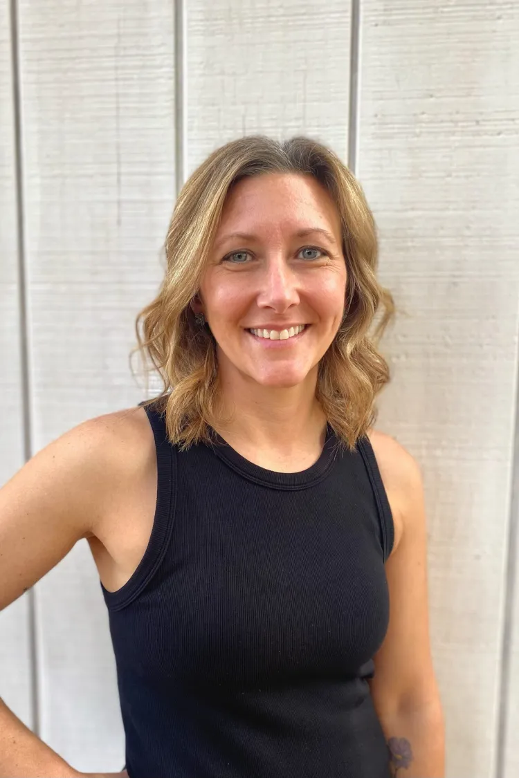 Smiling woman with wavy blonde hair in a black sleeveless top standing in front of a light wooden wall.